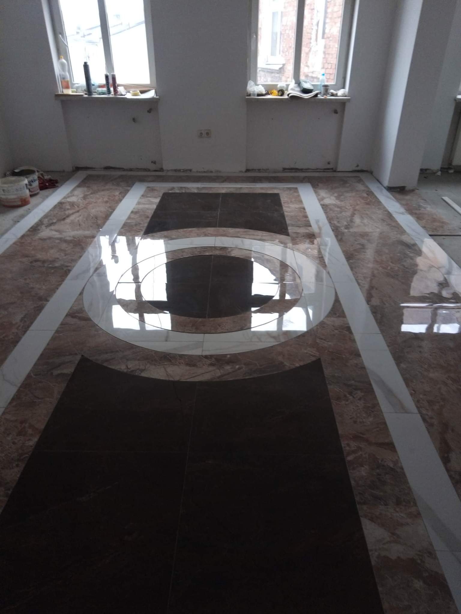 Nowoczesna podłoga z płytek w geometryczny wzór: brązowe płytki imitujące marmur z białymi wstawkami tworzącymi okrąg i prostokąty na tle surowych ścian i okien.