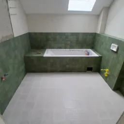 Wnętrze łazienki w Trzcianie w trakcie prac wykończeniowych, z zielonymi płytkami na ścianach do połowy wysokości i jasnymi płytkami na podłodze. Widoczna wanna obudowana płytkami, przygotowane...