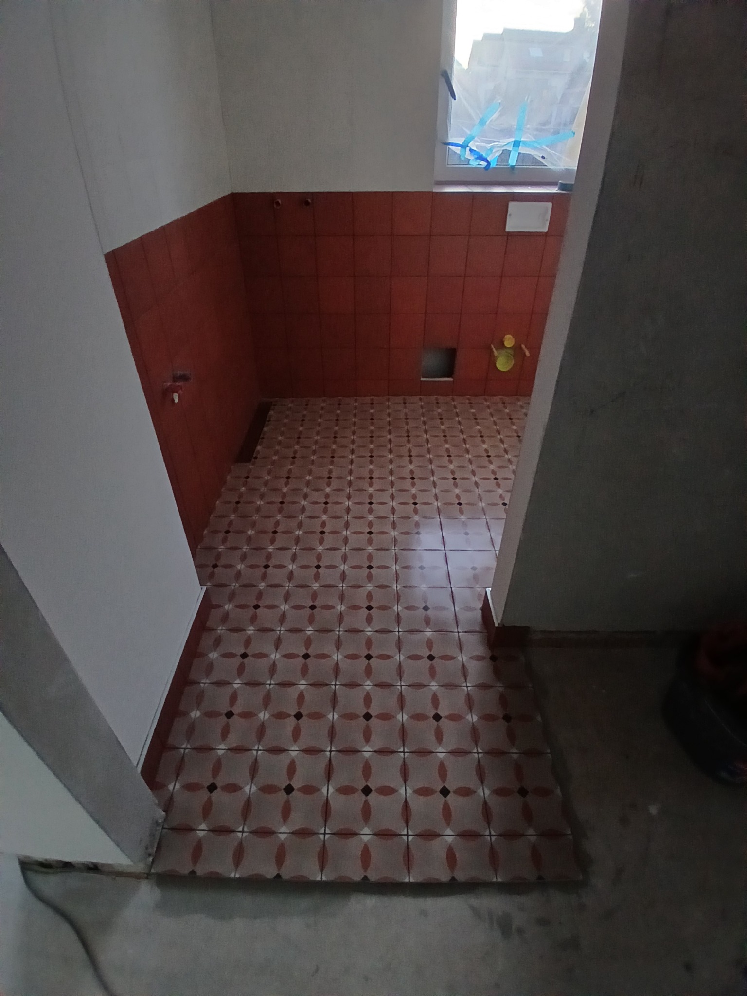 Wnętrze łazienki w trakcie remontu: widoczne pomarańczowe płytki na ścianach i podłodze z wzorem, zabezpieczone okno folią, surowy beton na podłodze przy wejściu.