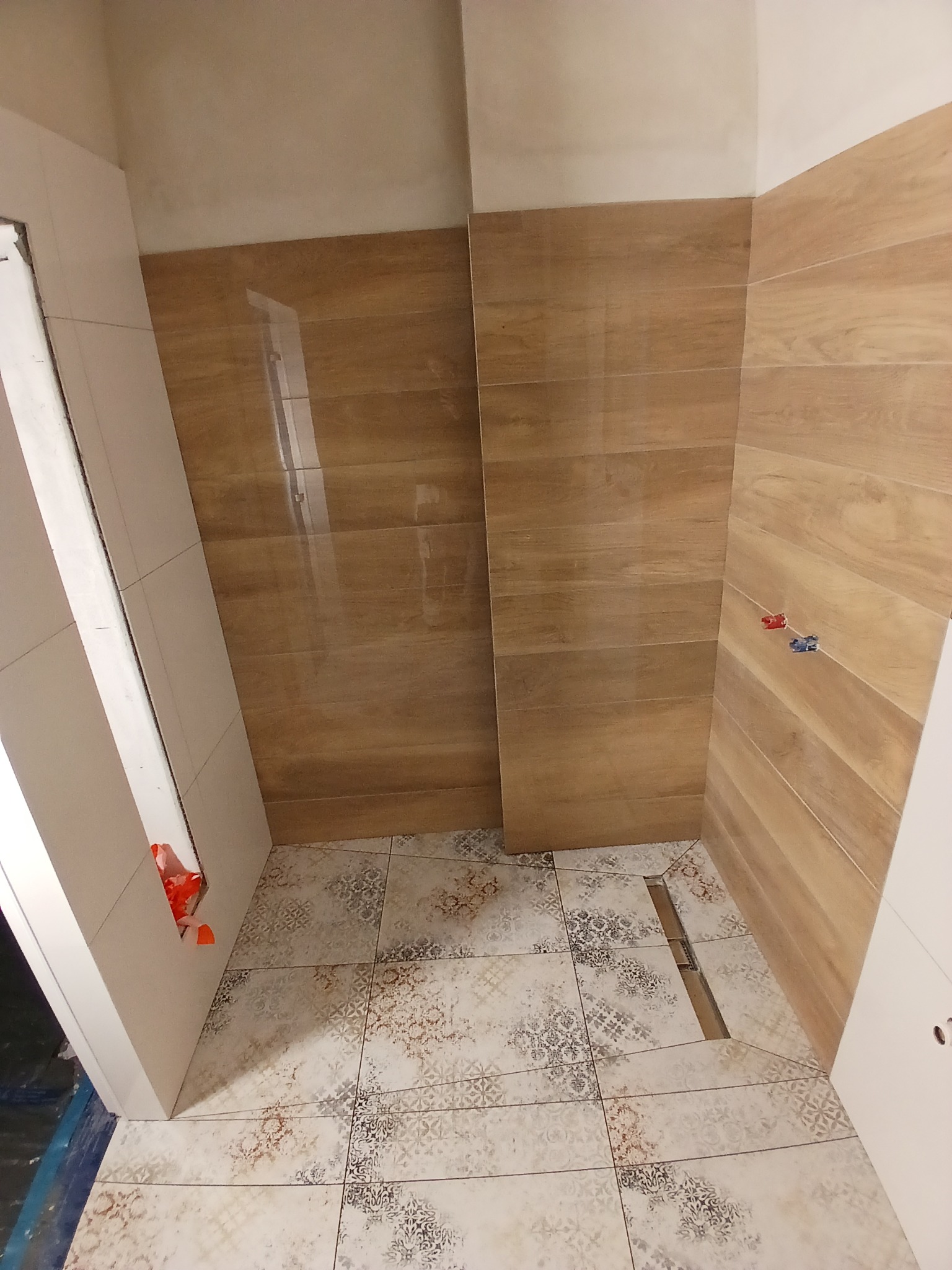 Wykończona łazienka z drewnopodobnymi płytkami na ścianach i wzorzystą podłogą. Widoczny odpływ liniowy i fragment drzwi z pomarańczowymi zabezpieczeniami.