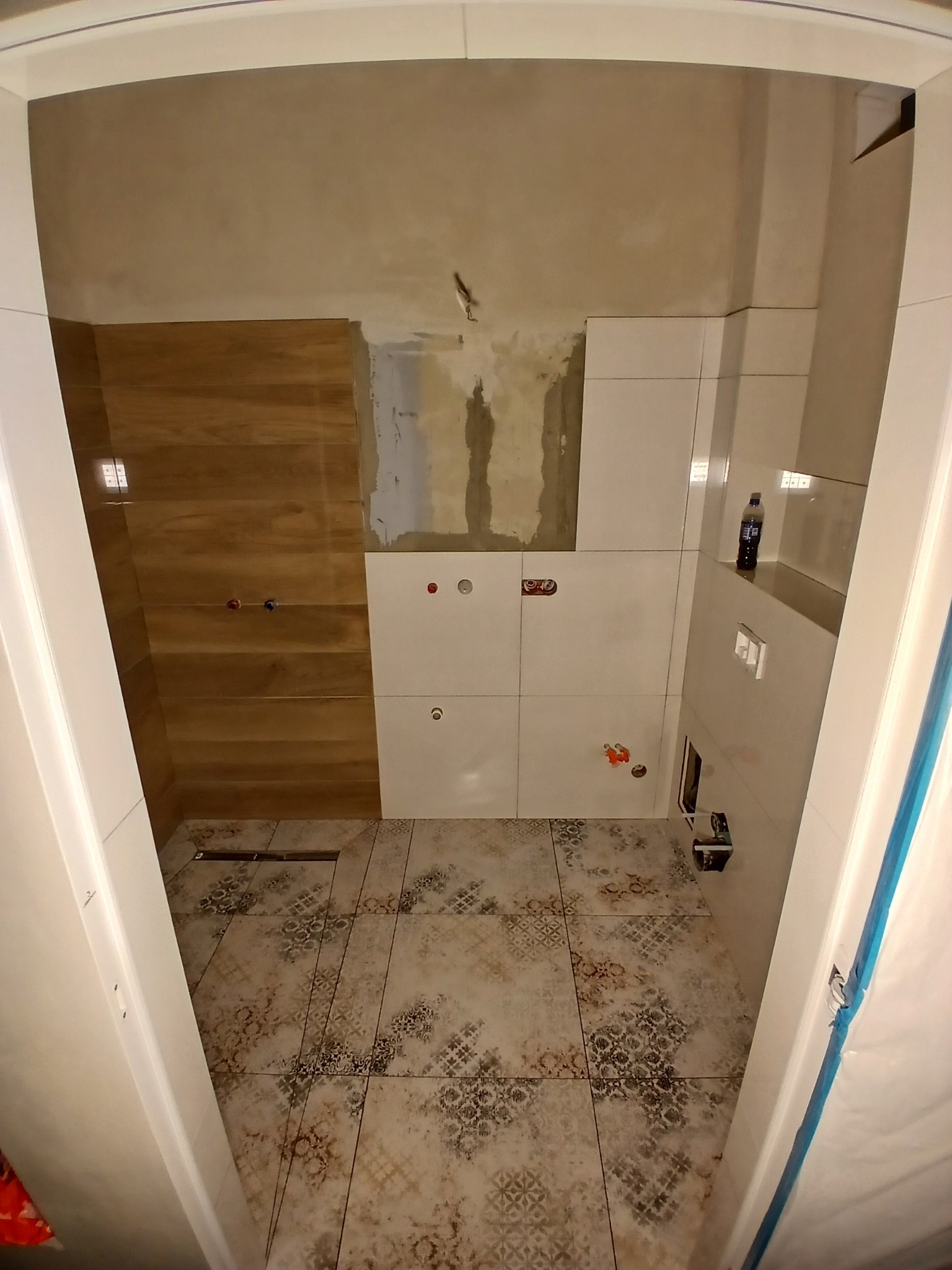 Wnętrze łazienki w trakcie remontu. Widoczne płytki imitujące drewno na ścianie, wzorzyste płytki podłogowe, częściowo położone białe płytki ścienne. Otwór drzwiowy z białym obramowaniem.
