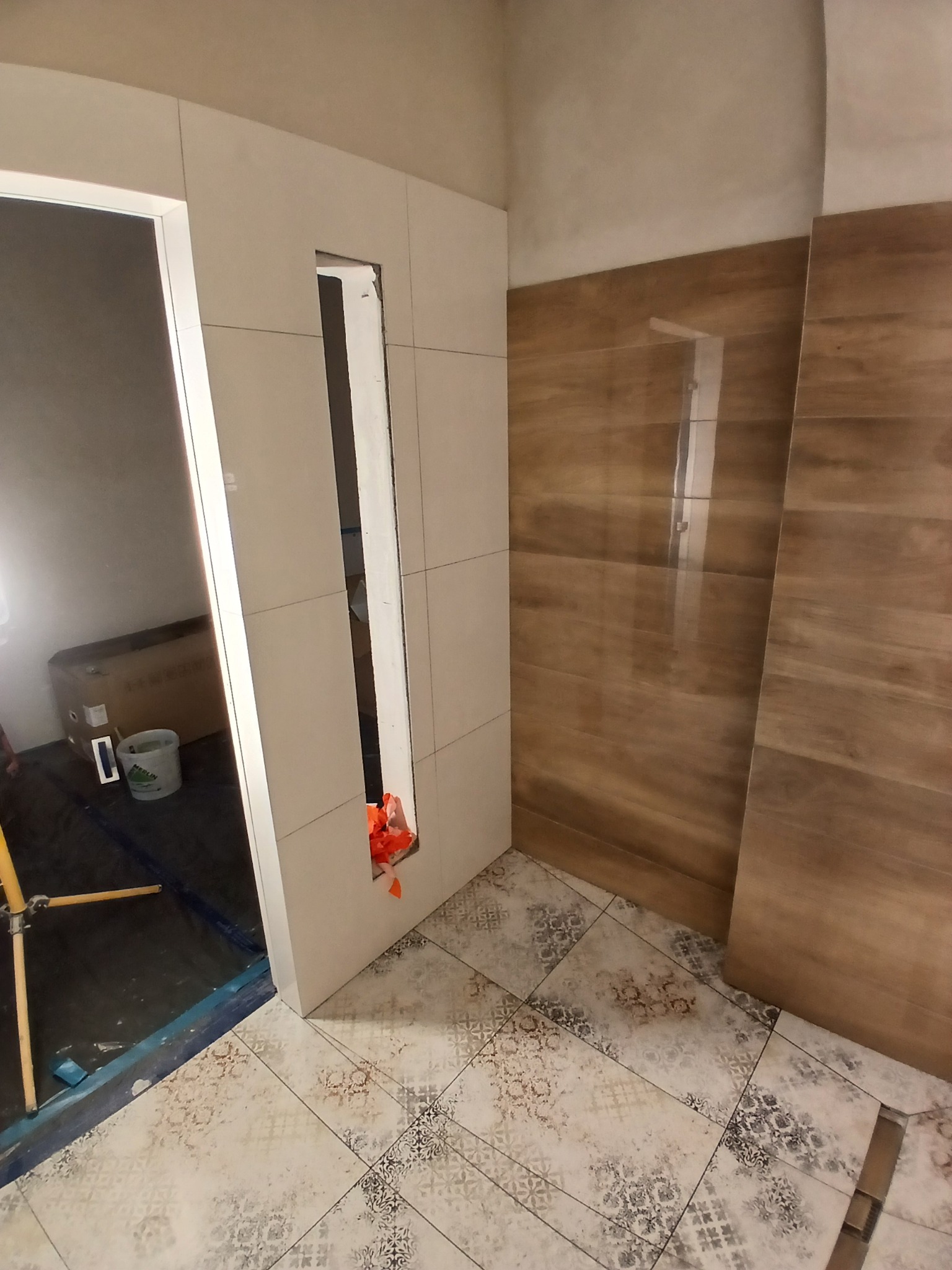 Układanie płytek w łazience: beżowe płytki ścienne z otworem na wnękę, drewnopodobne płytki i wzorzysta podłoga. Widoczne narzędzia i materiały budowlane.