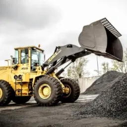 Żółta ładowarka kołowa marki XCMG z podniesioną łyżką nad stertą czarnego węgla na tle pochmurnego nieba.