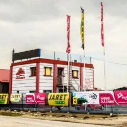 Budynek składu opału Adamex z banerami reklamowymi Jaret i Tauron, trzema flagami i stertą węgla w tle.