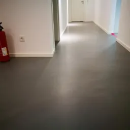 Szara, gładka podłoga w korytarzu, widoczny czerwony gaśnica przy ścianie i białe listwy przypodłogowe.