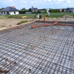 Zbrojenie fundamentu domu jednorodzinnego z pomarańczowymi rurami instalacji wodnej, widok z góry na siatkę stalową i betonową płytę.