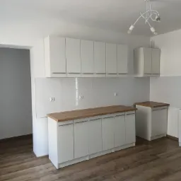 Jasna kuchnia z białymi szafkami i drewnianym blatem, widok na podłogę z paneli imitujących drewno i nowoczesną lampę sufitową.