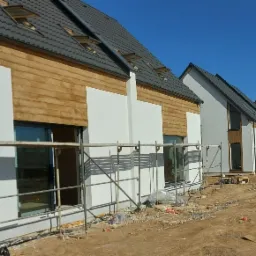 Szereg nowo wybudowanych domów jednorodzinnych z częściowo wykończoną elewacją: biały tynk i drewniana okładzina, widoczne rusztowania i teren budowy.