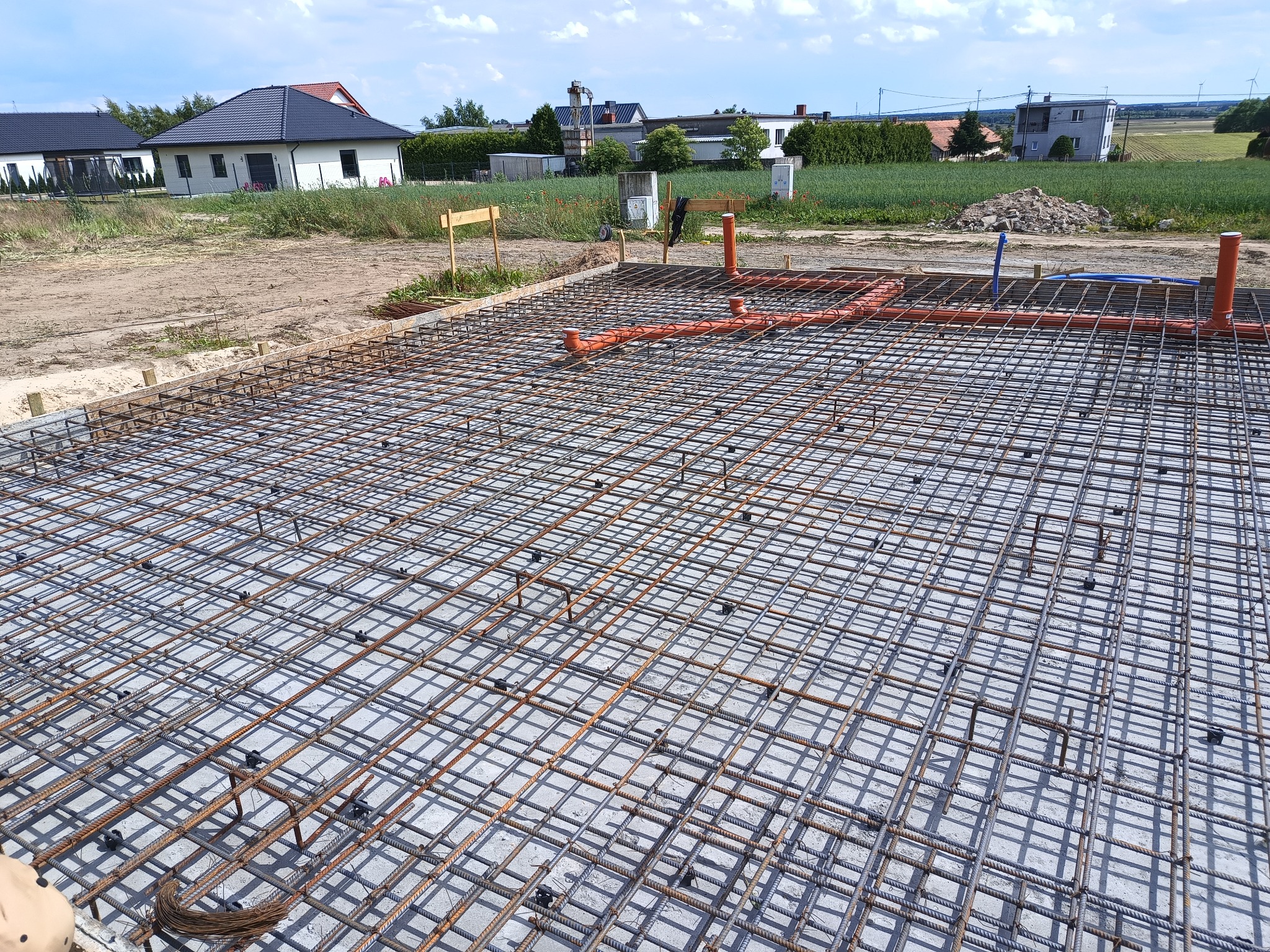 Zbrojenie fundamentu domu jednorodzinnego z pomarańczowymi rurami instalacji wodnej, widok z góry na siatkę stalową i betonową płytę.
