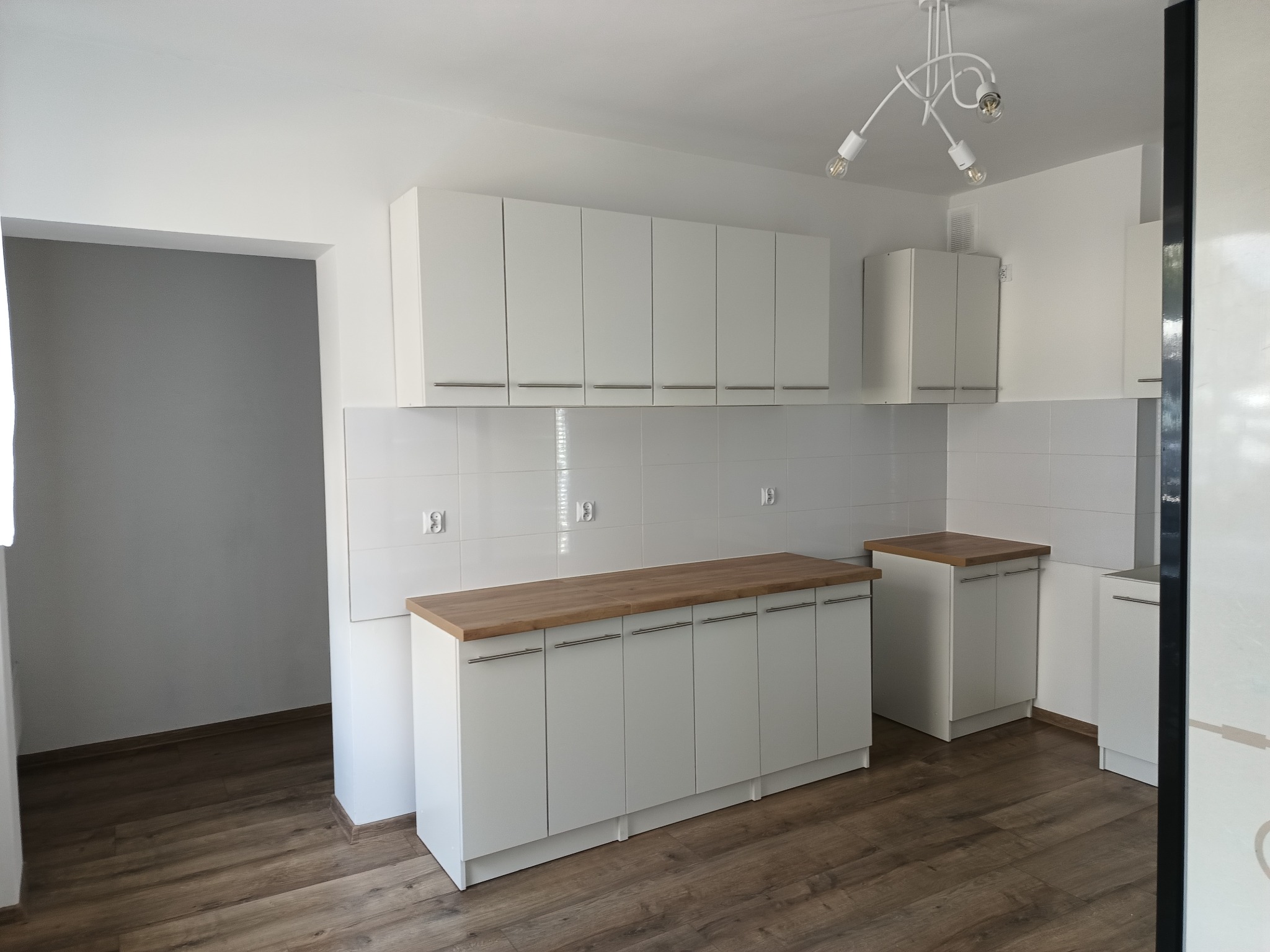 Jasna kuchnia z białymi szafkami i drewnianym blatem, widok na podłogę z paneli imitujących drewno i nowoczesną lampę sufitową.