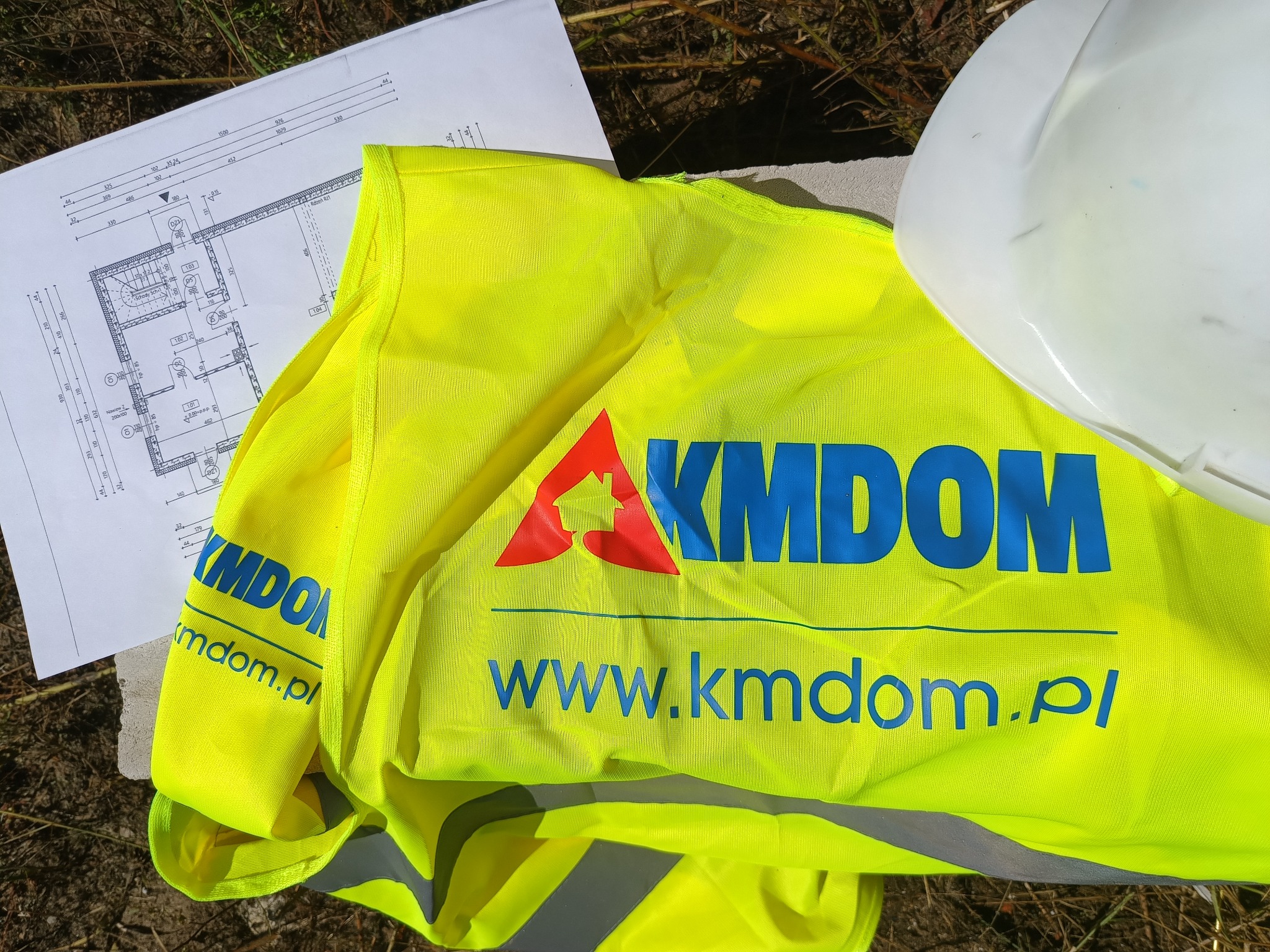 Plan architektoniczny domu, biały kask budowlany i żółta kamizelka odblaskowa z logo firmy KMDOM leżą na ziemi.