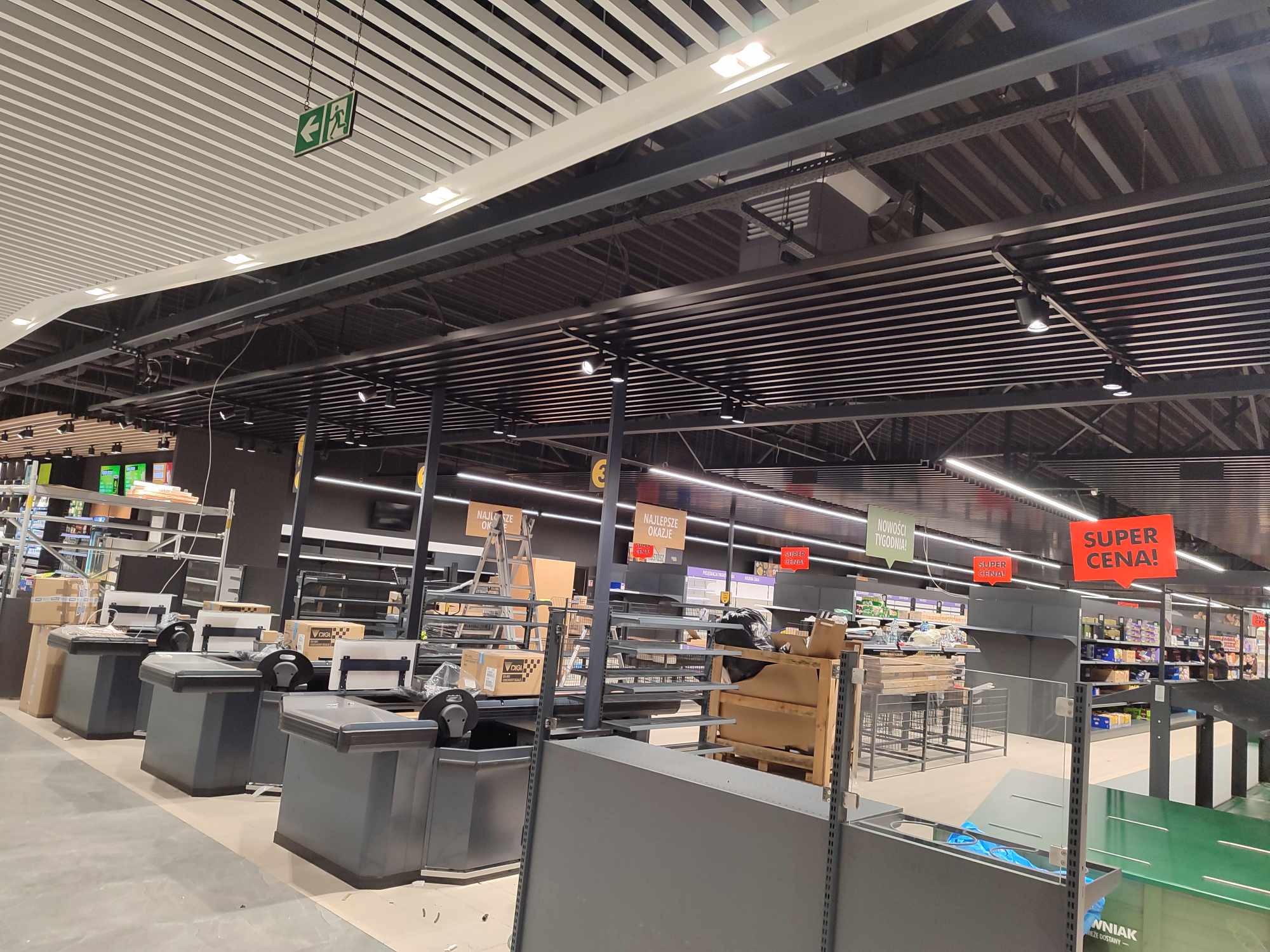 Wnętrze sklepu w trakcie remontu lub aranżacji: widoczne kasy, regały, drabina, elementy konstrukcyjne sufitu i oświetlenie, z częściowo rozpakowanym towarem i elementami wyposażenia.