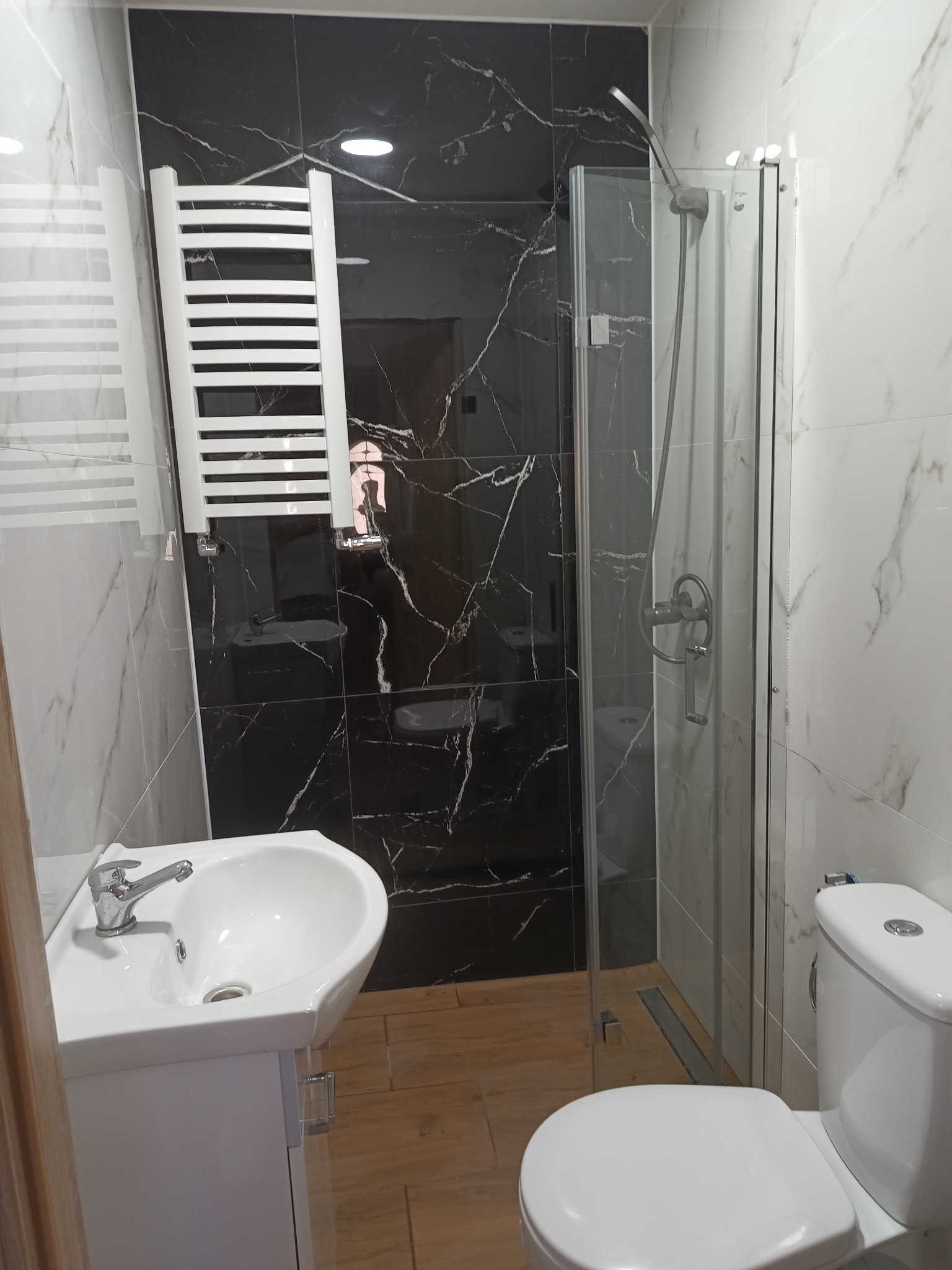 Nowoczesna łazienka z czarno-białymi marmurkowymi płytkami, kabiną prysznicową ze szklanymi drzwiami, umywalką z szafką i grzejnikiem drabinkowym.