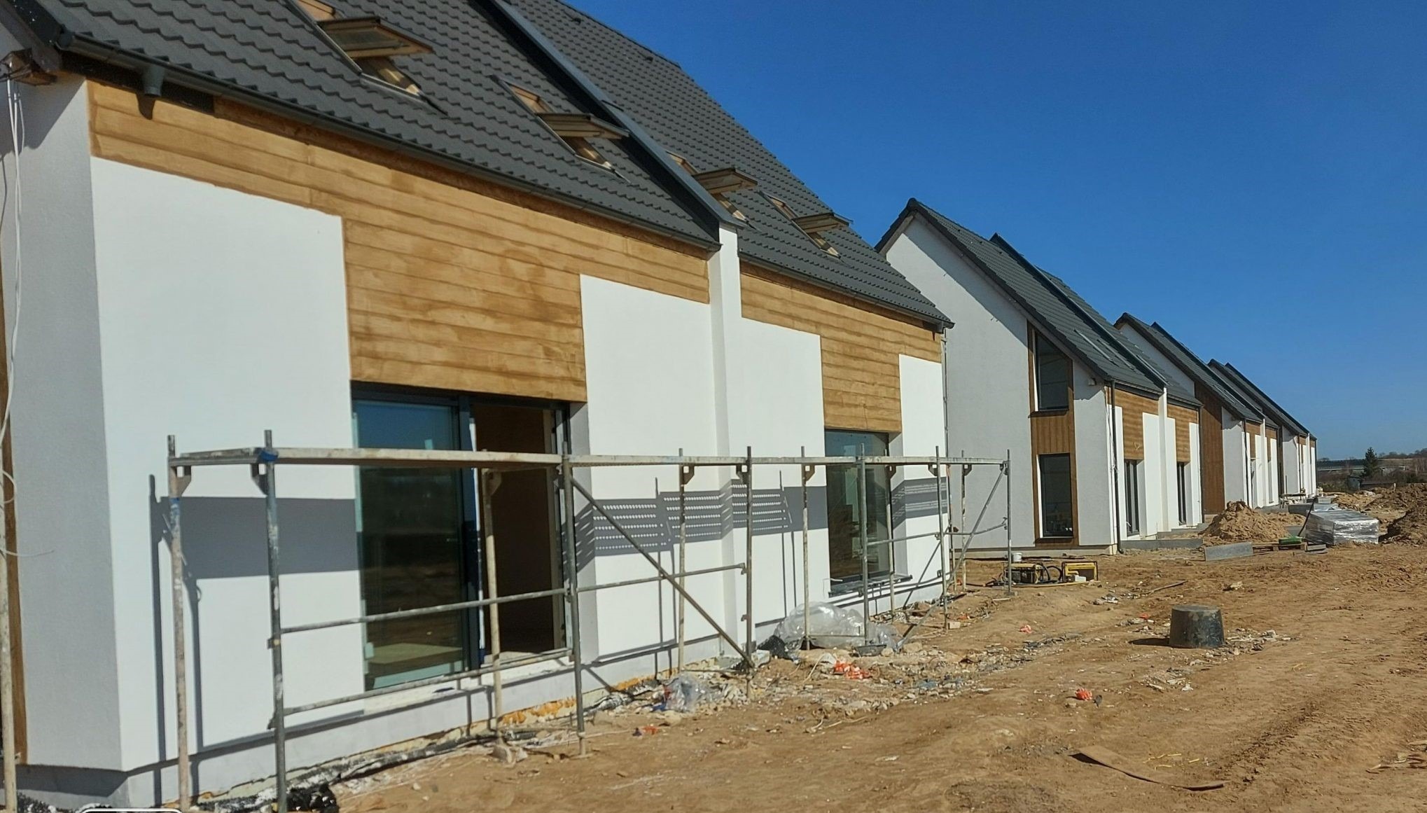 Szereg nowo wybudowanych domów jednorodzinnych z częściowo wykończoną elewacją: biały tynk i drewniana okładzina, widoczne rusztowania i teren budowy.