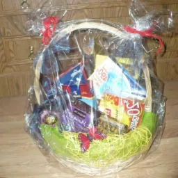 Wypełniony słodyczami wiklinowy kosz, owinięty przezroczystą folią i przewiązany czerwoną wstążką, z dekoracją w postaci sztucznego motyla.