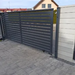 Nowoczesna, przesuwna brama wjazdowa z poziomymi panelami w kolorze grafitowym, częściowo otwarta, z widocznym napędem i betonowym słupkiem obok, na tle kostki brukowej i zabudowy jednorodzinnej.
