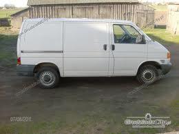 Biały samochód dostawczy (van) marki Volkswagen Transporter T4 na trawiastym podłożu, z widocznym budynkiem gospodarczym w tle. Zdjęcie zrobione z niskiej perspektywy.