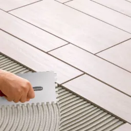 Układanie podłużnych, jasnych płytek podłogowych z imitacją drewna, widoczna ręka glazurnika w czerwonej koszuli w kratę rozprowadzająca klej za pomocą pacy ząbkowanej.