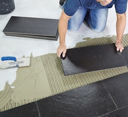 Układanie ciemnych płytek podłogowych na warstwie kleju, widoczna paca z ząbkami i stos płytek w tle.