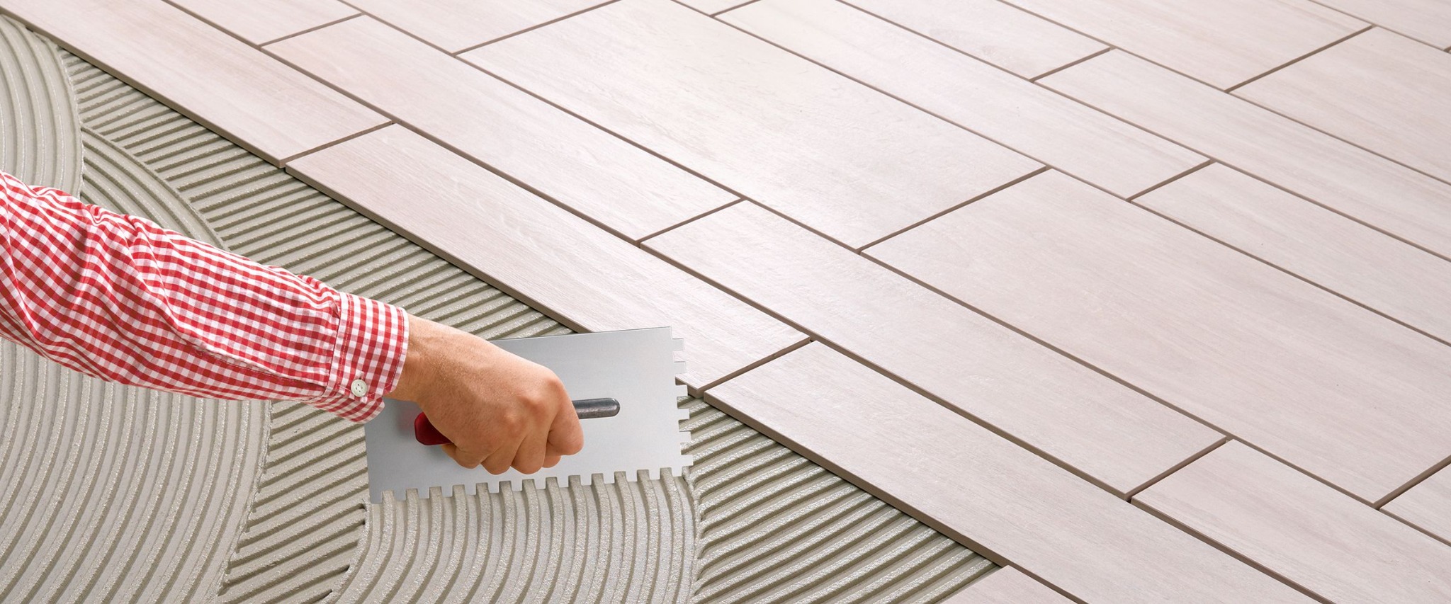 Układanie podłużnych, jasnych płytek podłogowych z imitacją drewna, widoczna ręka glazurnika w czerwonej koszuli w kratę rozprowadzająca klej za pomocą pacy ząbkowanej.