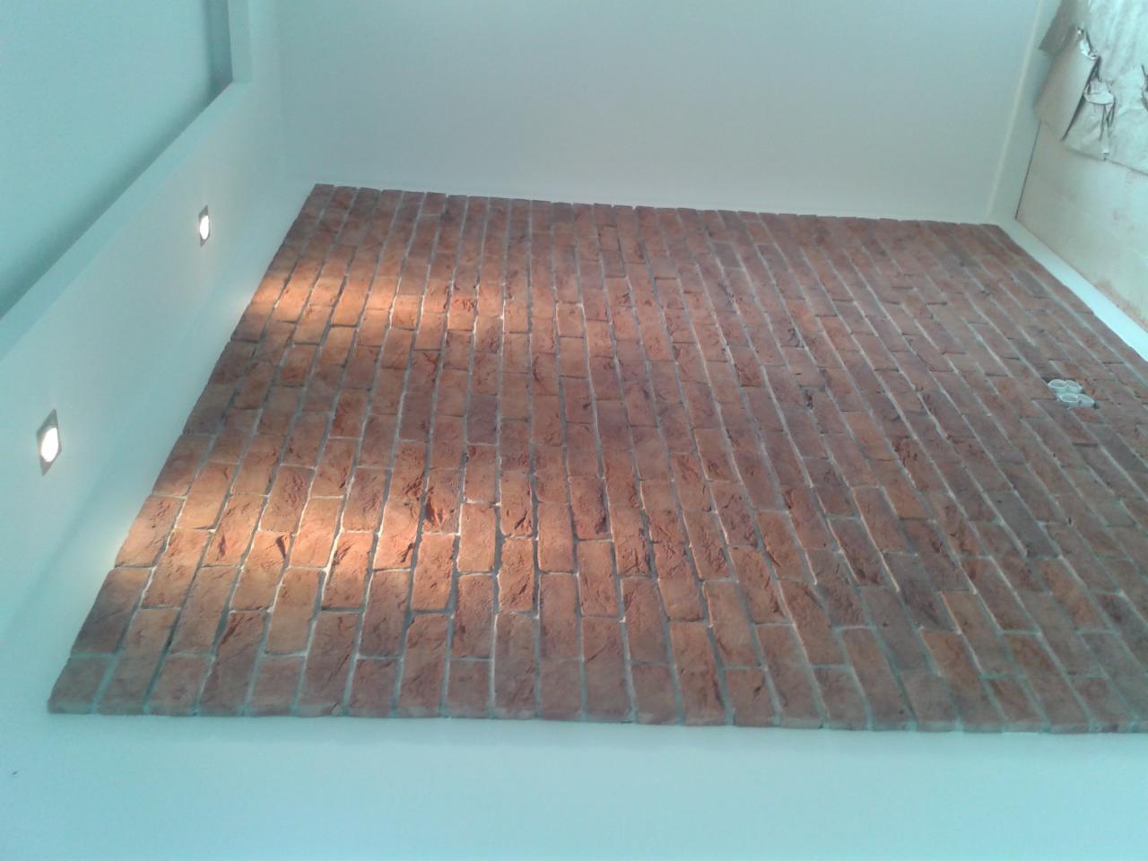 Ściana wykończona cegłą klinkierową w pomieszczeniu z białymi ścianami i wbudowanymi punktowymi lampami oświetleniowymi.