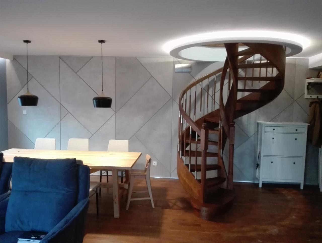 Jasne wnętrze z drewnianymi schodami spiralnymi, geometryczną ścianą akcentującą, drewnianym stołem jadalnym i trzema wiszącymi lampami z czarnymi kloszami.