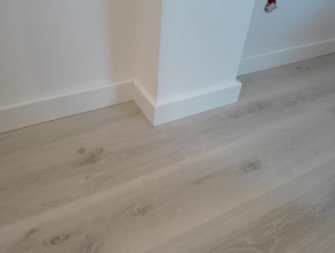 Jasne panele podłogowe ułożone przy białej ścianie z listwami przypodłogowymi, widoczny fragment narożnika ściany i gniazdko elektryczne.