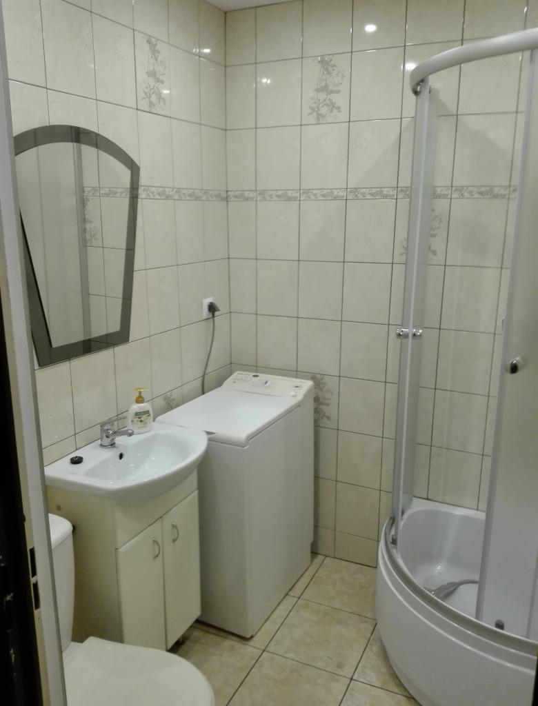 Jasna łazienka po remoncie: kabina prysznicowa z zaokrąglonym frontem, umywalka z szafką, pralka i lustro w dekoracyjnych płytkach.