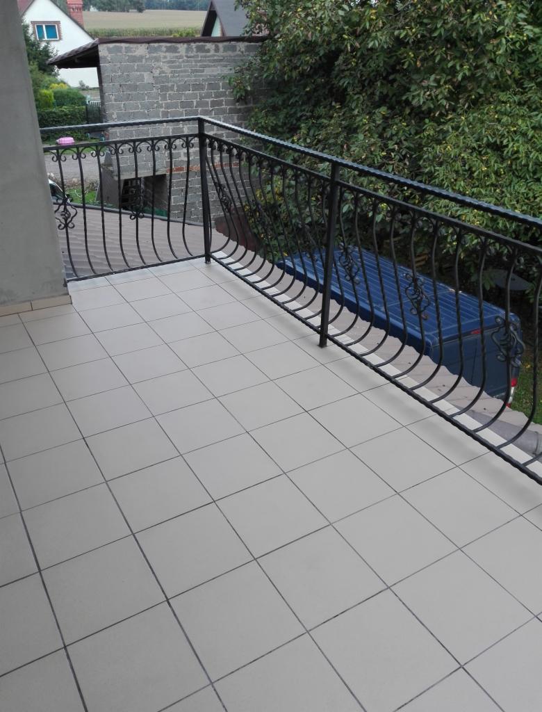 Balkon wyłożony jasnymi, kwadratowymi płytkami ceramicznymi, z czarną, metalową balustradą o dekoracyjnych, zakrzywionych elementach. W tle widoczny budynek z szarej cegły i zieleń drzew.