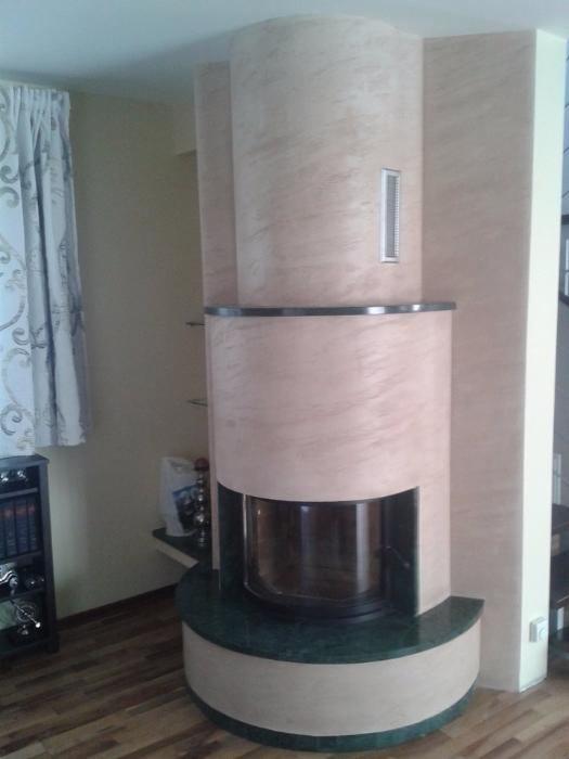 Otynkowany kominek w odcieniach beżu i zieleni, z widocznym paleniskiem i dekoracyjnymi półkami w tle, na drewnianej podłodze.