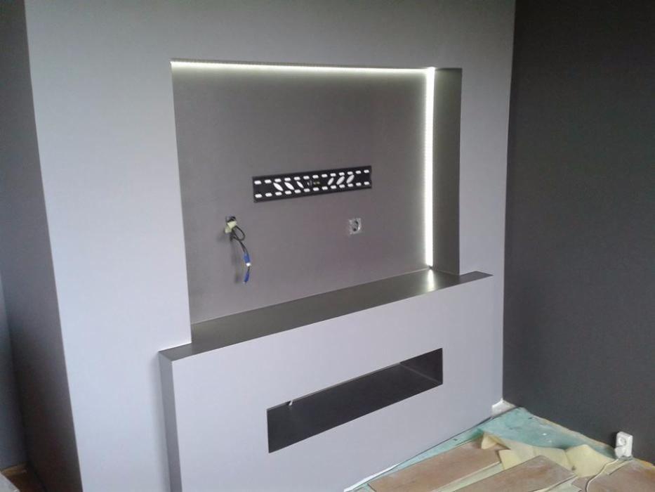 Szara ściana z wnęką pod telewizor z podświetleniem LED, widoczne kable i uchwyt montażowy, fragment podłogi z panelami.