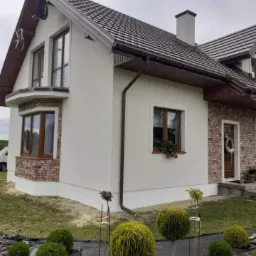 Dom jednorodzinny z ocieploną, białą elewacją, fragmentem wykończenia z cegły klinkierowej i brązowymi ramami okiennymi, widoczne krzewy ozdobne w ogrodzie.