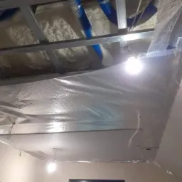 Poddasze w trakcie izolacji pianą, widoczne niebieskie rury wentylacyjne, metalowa konstrukcja sufitu, folia paroizolacyjna i fragment pokoju z oknem.