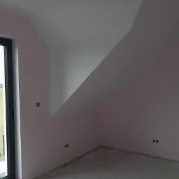 Poddasze w trakcie wykańczania, ściany pomalowane na jasny róż, surowa wylewka na podłodze, widoczne gniazdka elektryczne i puszka instalacyjna, okno z widokiem na zielony krajobraz.