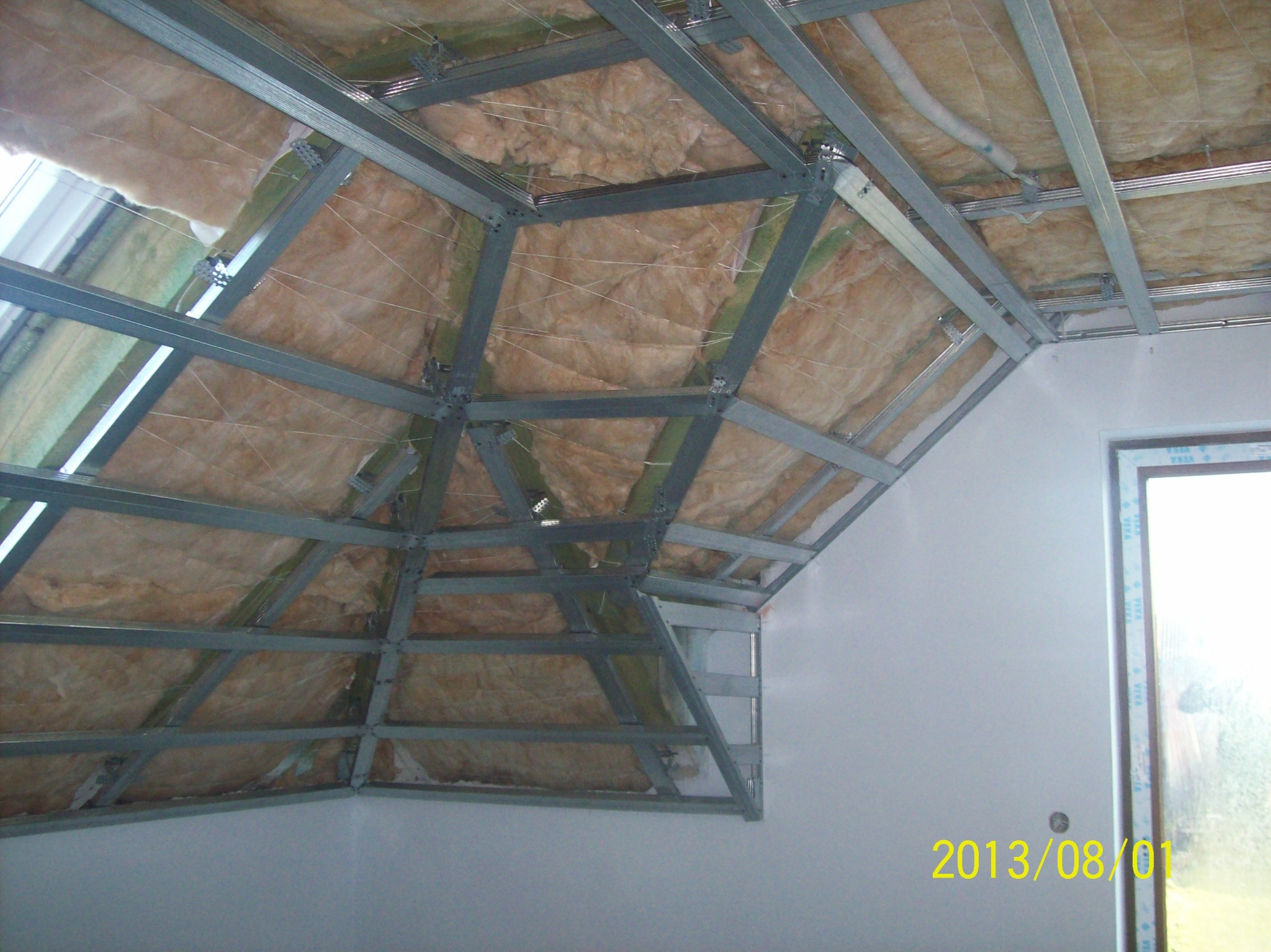 Skośny dach poddasza w trakcie ocieplania, widoczne wełna mineralna, metalowy stelaż i fragment okna dachowego, data na ścianie 2013/08/01.