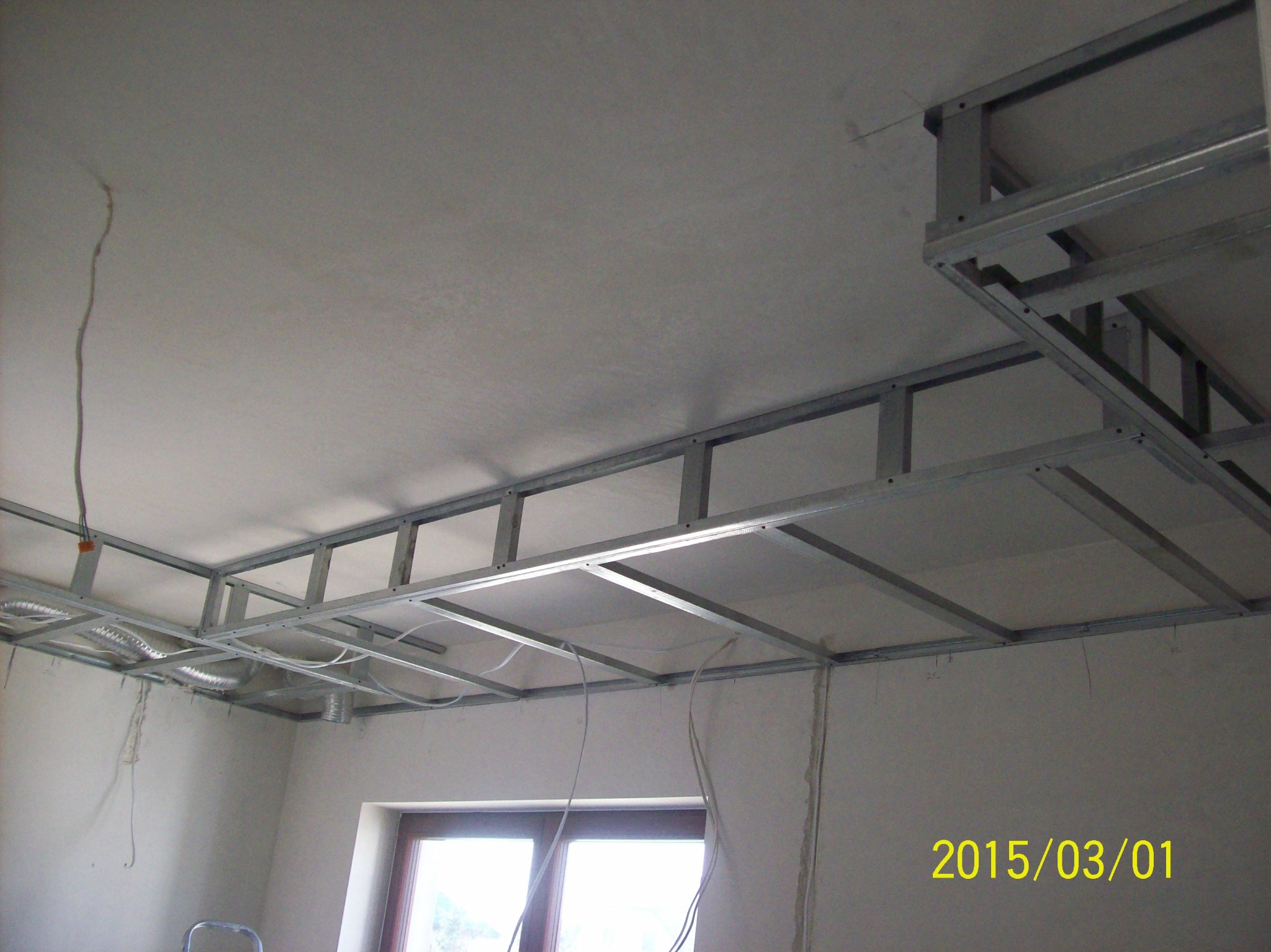 Montaż metalowej konstrukcji sufitu podwieszanego z płyt kartonowo-gipsowych, widoczne przewody i okno w tle.