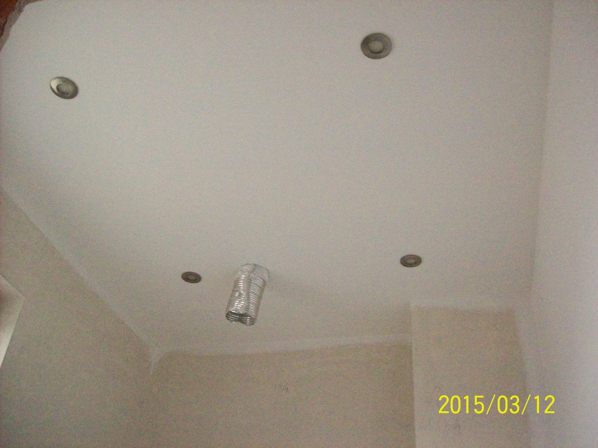 Biały sufit z czterema okrągłymi lampami halogenowymi i wystającym fragmentem srebrnej, elastycznej rury wentylacyjnej w trakcie prac wykończeniowych.