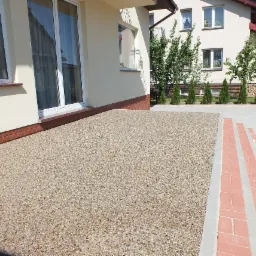 Nawierzchnia mineralno-żywiczna, wodoprzepuszczalna, ułożona przed budynkiem z oknami PCV i ceglaną podmurówką, widok z poziomu gruntu na fragment posesji.