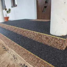 Dwustopniowe schody wejściowe do domu wykończone czarną i brązową nawierzchnią mineralno-żywiczną z delikatnym złotym obramowaniem, otaczające białą kolumnę przy drzwiach wejściowych.