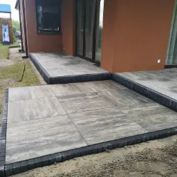 Ułożony taras z szarych płyt betonowych obramowany ciemnoszarą kostką, widok na brązową elewację domu z oknami i plac zabaw w tle.