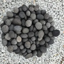 Czarny, porowaty kamień dekoracyjny w metalowym koszu na tle drobnego, białego kruszywa.