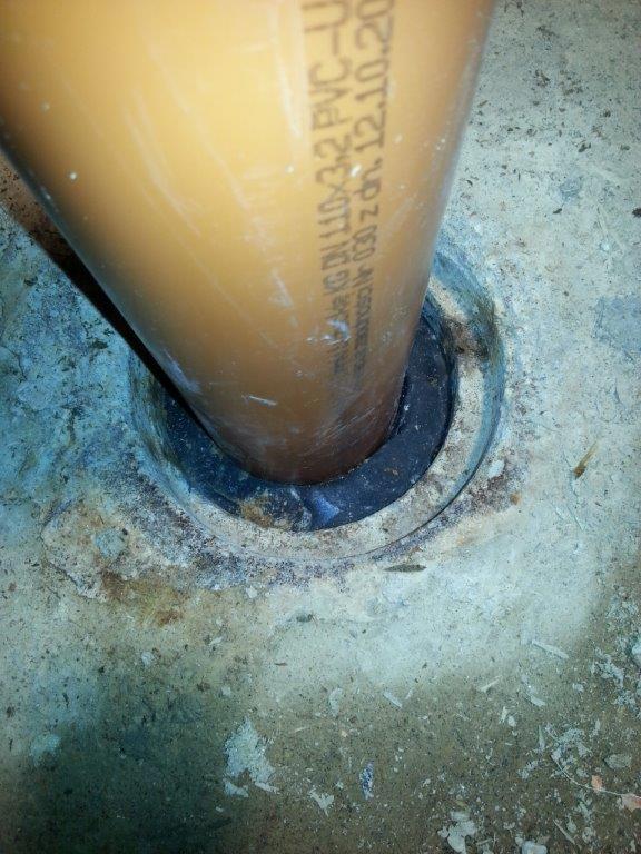 Brązowa rura kanalizacyjna PVC uszczelniona czarnym materiałem w betonowej posadzce, widoczne oznaczenia na rurze.