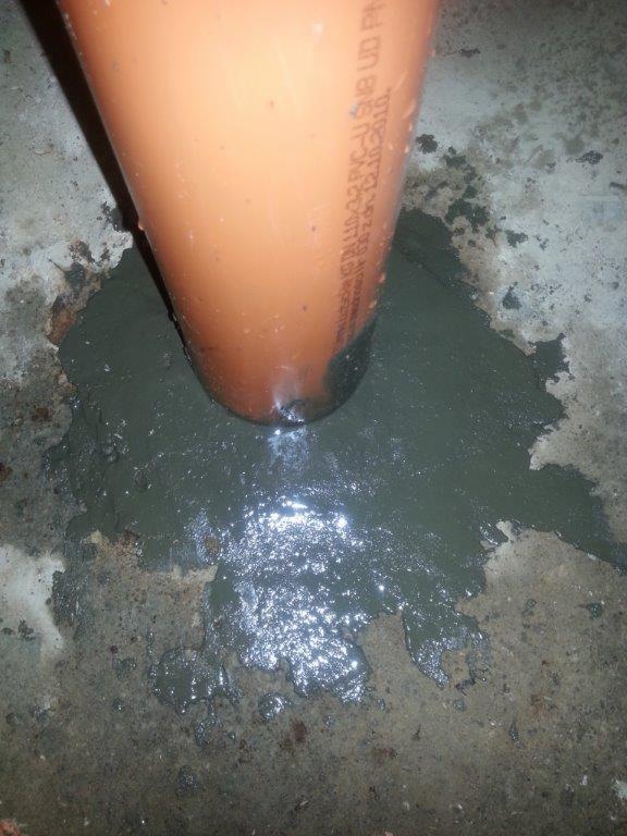 Pomarańczowa rura PVC uszczelniana szarym uszczelniaczem na betonowej powierzchni podłoża. Widoczne napisy na rurze.