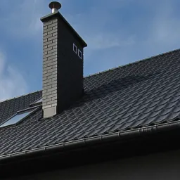 Czarny dach z dachówki ceramicznej z kominem z cegły i systemem rynnowym, widok z dołu na tle błękitnego nieba z chmurami.