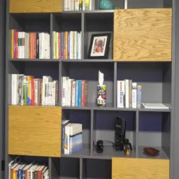 Szara, modułowa biblioteka z drewnianymi wstawkami, wypełniona książkami, zdjęciem w ramce, ceramiczną figurką ptaka oraz mikrofonem na statywie.