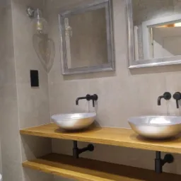 Dwie srebrne umywalki w kształcie misy z czarnymi bateriami, zamontowane na drewnianym blacie z półką, szare płytki na ścianie i dwa lustra w srebrnych ramach.