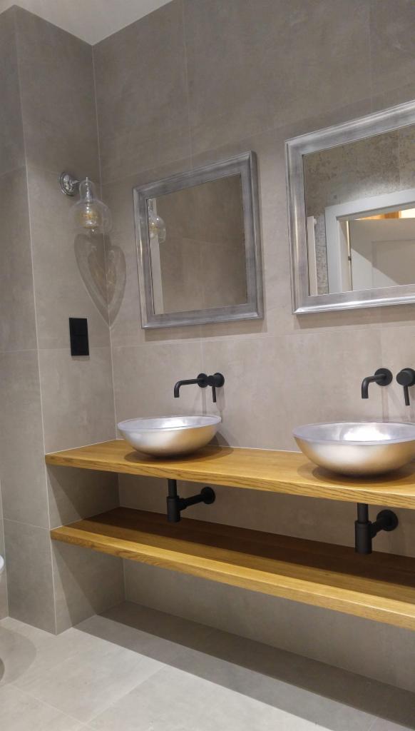 Dwie srebrne umywalki w kształcie misy z czarnymi bateriami, zamontowane na drewnianym blacie z półką, szare płytki na ścianie i dwa lustra w srebrnych ramach.