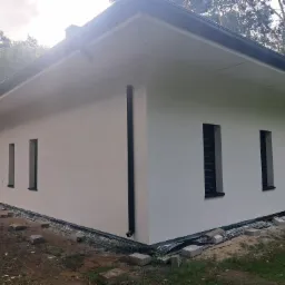 Biały, parterowy dom z czarnymi akcentami (rynny, okna). Wykończona elewacja z widoczną izolacją fundamentów i elementami krajobrazu. Dom otoczony drzewami.