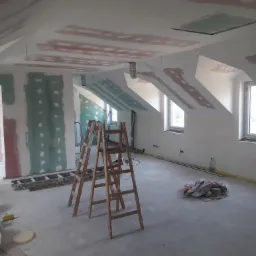 Poddasze w trakcie prac wykończeniowych: widoczne płyty kartonowo-gipsowe na ścianach i skosach, częściowo zaszpachlowane, drewniana drabina, narzędzia i materiały budowlane na podłodze, okna...