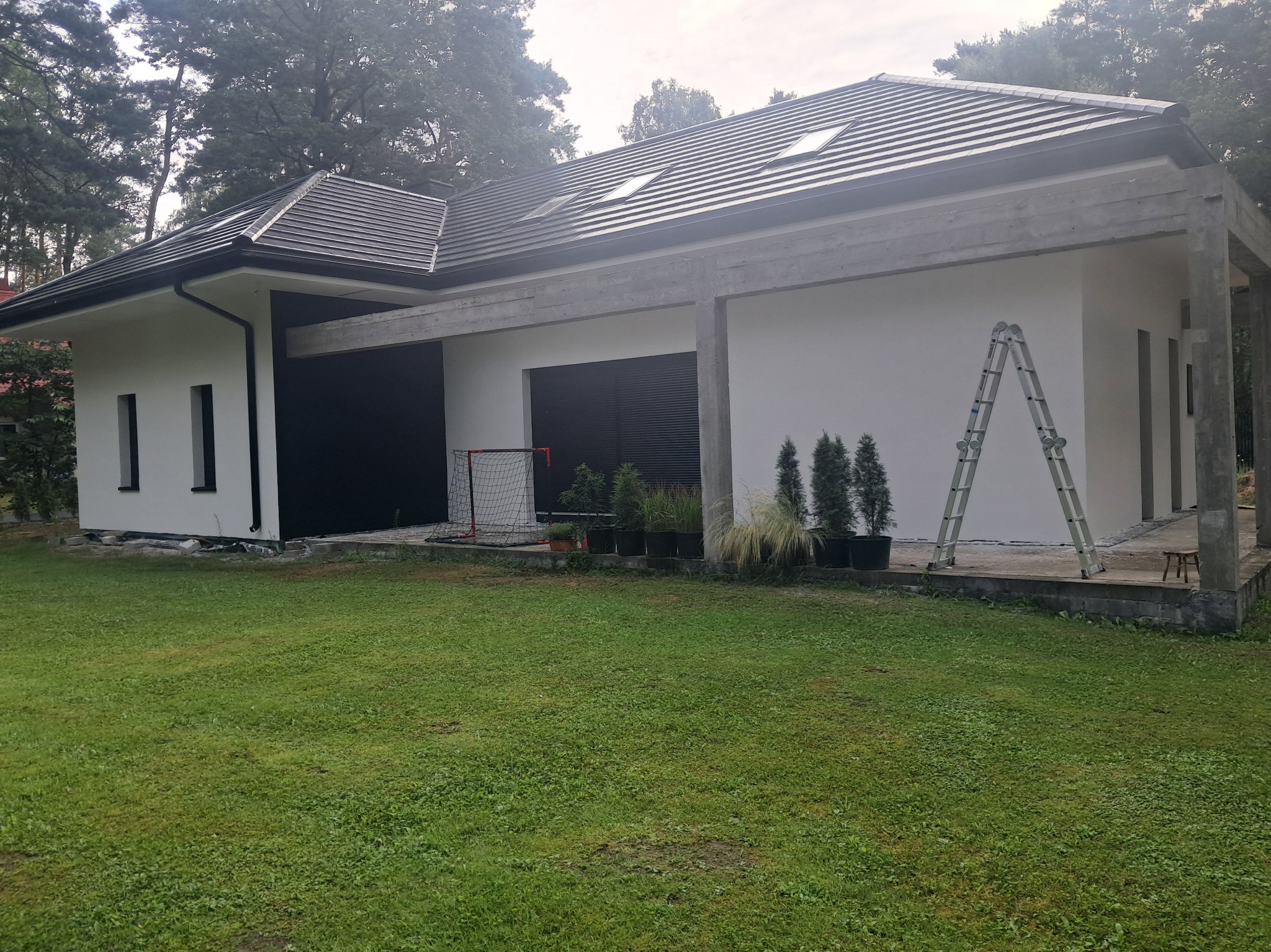 Biały dom z szarą konstrukcją i ciemną stolarką. Na trawniku drabina, donice z roślinami i bramka do piłki nożnej. Widok z zewnątrz, w tle drzewa.